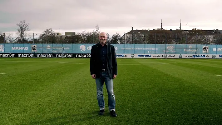 Bewerbungsvideo 1 Mann im dunklen Sakko steht auf dem Rasen eines Fußball-Trainingsplatzes.