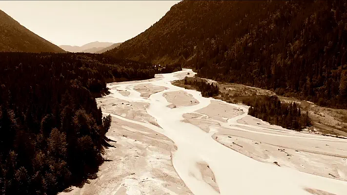 Drohnenaufnahmen 1 Drohnenaufnahme eines verzweigten Flussbetts in einem bewaldeten Bergtal, in Sepiatönen.