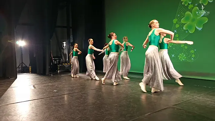 Making Of Video 1 Dancers in green leotards and long skirts rehearsing on a stage in front of a green wall.