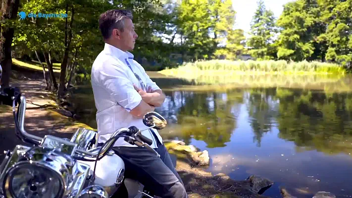 Recruiting Film 5 Man in a white shirt leaning with folded arms on a motorcycle by a lake.
