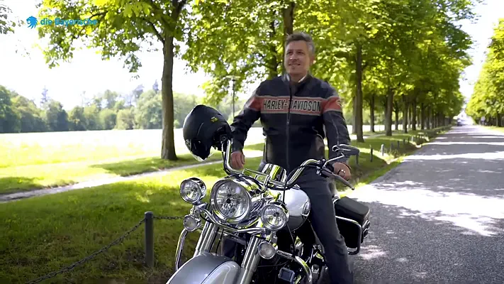 Recruiting Film 4 Motorcyclist in a leather jacket holding his helmet next to his bike in a park.