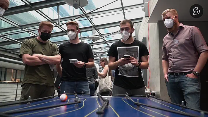 Schulungsvideo 3 Junge Männer mit Masken verfolgen ein Geschicklichkeitsspiel an einem Messestand.